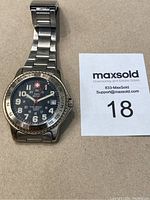 Front view of Swiss Military wristwatch with black dial, large numerals, red logo, date window and silver tachymeter bezel on stainless steel bracelet.