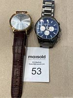 Two gents watches side by side on a neutral surface with a MaxSold lot number card.