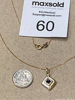 Pendant with square sapphire center, two small diamonds, and gold chain next to a quarter for size reference.