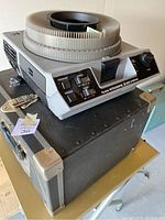 Kodak Ektagraphic III AMT slide projector with circular carousel slide tray mounted on top, placed on its black carrying case with metal reinforced corners.