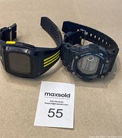 Two sports watches on a flat surface with a MaxSold lot card '55'. One is Casio G-Shock, and the other is Adidas.