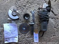 Top-down photo showing angle grinder with coiled power cord, protective metal guard, wire brush attachment, diamond cutting disc, wrench, and instruction manual on concrete floor.