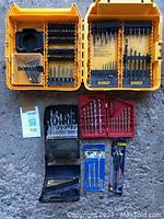 Top view of a yellow DEWALT plastic case open with assorted screw and drill bits arranged in compartments. Below are additional bit sets and packs including black and red cases, and small blue and black packages containing drill bits.