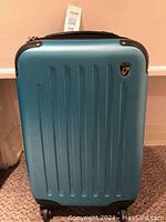 Front view of blue hard shell carry on suitcase with vertical ridges and Hyes brand logo badge on the front.