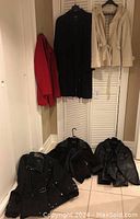Photo of six ladies coats and jackets hanging and lying on floor showing variety of styles and colors including red, cream, and multiple blacks.