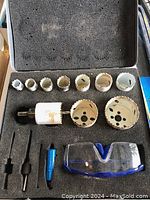 Open metal case with foam insert displaying 7 white hole saw blades, 2 black mandrels, a pilot drill bit, a blue accessory tool, and clear safety goggles with blue trim.
