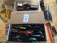 Overview of various hand tools including screwdrivers, chisels, measuring tapes, and a hand saw arranged in plastic bins and a cardboard box on a wooden surface.
