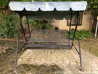 Front view of the black iron mesh porch swing with blue fabric canopy, showing overall structure and condition on a stone patio.