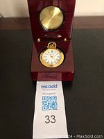 Pocket watch resting inside open wooden presentation box showing face and circular brass accent inside lid