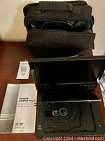 Sony portable DVD player open showing LCD screen, disc tray with laser lens, user manual beside it, and black carrying case behind.
