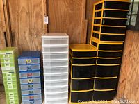 Wide shot showing all five plastic storage drawer units in various colors and sizes including green, blue, white translucent, and black with yellow trim against wooden wall.