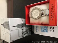 Silver-tone Caravelle New York wristwatch with rhinestone bezel in original red display case and white outer box featuring Empire State Building image.