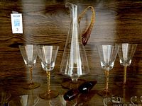 Front view showing six clear wine glasses with amber twisted stems and bases arranged around a tall clear glass decanter with an amber twisted handle on a wooden surface.