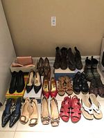 Wide view of 13 pairs of women's shoes arranged on the floor: includes boots, heels, sandals, and one box with matching dress shoes and purse.
