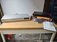 Photo shows silver Aiwa CXD-AX1 DVD/CD player on left and black Sony DVP-SR20 DVD player on right with remote, box, and instructions on wooden table.