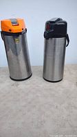 Two stainless steel coffee airpot dispensers, one with orange plastic top, the other with black top and handle, standing side by side on a surface