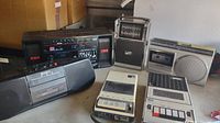 Five vintage electronic audio devices placed on a table, visible from front and side angles showing brand logos, cassette windows, buttons and speaker grilles.
