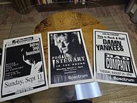 Three vintage-style rock concert posters on a wooden table: Rod Stewart, Roger Daltrey with Pete Townshend, Damn Yankees and Bad Company.