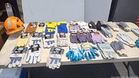 Wide shot of table with over 30 pairs of assorted new work gloves in various colors and styles. Orange safety hat visible but not included in sale.