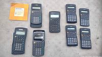 Eight scientific calculators arranged on a fabric surface. Includes multiple Texas Instruments and unknown brand calculators plus a small pamphlet.