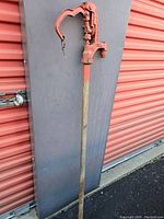 Full length view of red cast iron hand water pump showing handle, spout, and long cylindrical base against a grey backdrop with red door.
