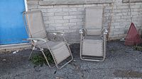 Two beige fabric sling chaise lounge chairs with metal frames positioned outdoors against a brick wall and asphalt ground.
