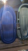 Two large plastic sleds side by side outdoors, one blue and one gray, showing overall shape and handle placements.