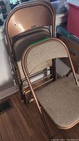 Three folding card chairs stacked with bronze metal frames and beige padded seats and backrests.