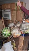 Photo showing nine large vintage-style ribbed glass Christmas ball ornaments in metallic silver, green, gold, and bronze colors with metal caps and attached green strings for hanging.