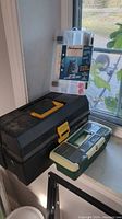 Two fishing tackle containers, one black and yellow box and two clear compartment containers, one containing fishing tackle and hooks.