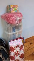 Image showing a stack of gift wrap bags, a large bag of pink decorative shred, and transparent plastic storage containers holding wrapping supplies against a white wall and wooden floor.