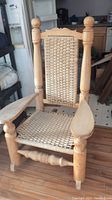 Photo of a large wooden rocking chair with woven cane seat and backrest. Chair has thick turned wood posts and wide arm rests. Floor and background shelving visible.