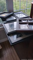 Photo showing six picture frames stacked on a table by a window. Frames vary in size and configuration including single, double, triple and multi-image openings. Wooden or wood-like materials with dark colors visible.