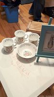 Photo of four Royal Albert Peter Rabbit mugs, one small bowl with Peter Rabbit design, and a small blue easel holding a framed Peter Rabbit picture, placed on a hardwood floor.