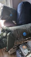 Photo showing three sleeping bags including two green bags with circular patch logos and one blue sleeping bag in compression sack. Brown blanket and a gray ground cover with orange label also visible.