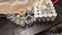 Photo showing multiple packs of tea light candles in various colors and several glass candle holders on a black surface.