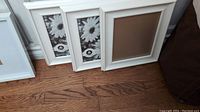 Five white picture frames leaning against a wall on hardwood floor, two have daisy placeholder pictures.