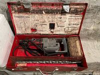 Milwaukee rotary hammer drill in red metal case with attached long auger drill bit and packaging paper inside case.