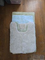 Three stacked bath mats of different colors and sizes: beige U-shaped, light blue rectangular, and green rectangular, showing material and size variation.