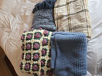 Four folded blankets and throws stacked on a bed including a crochet floral throw, blue waffle-textured throw, dark grey fringed throw, and beige plaid blanket.
