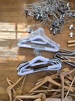 Photo showing a pile of silver metal hangers with clips, white plastic hangers with clear plastic covers, and some black hangers stacked together on a wooden floor.