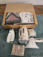 Photo showing all items laid out near an open cardboard box including two white handheld clothes steamers, iron, accessories with plastic wrapping, and instruction manuals.