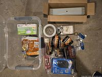 Top-down view of assorted household hardware and supplies including batteries, light fixtures, and small hardware in plastic container and loose packages.