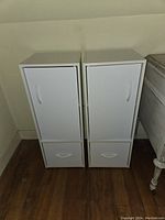 Two matching white cupboards placed side by side on a wooden floor against a wall, showing their front with closed doors and drawers.