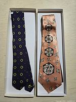 Two ties in white boxes on a flat surface: navy blue with yellow and white circular pattern and peach with black and green geometric floral pattern.