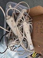 Photo showing a tangled collection of white power cords and two white power strips inside a plastic bin, along with a cardboard box labeled 'Power Cords'.