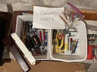 Photo shows two white bins filled with assorted small hand tools including screwdrivers, pliers, wire cutters, and a vintage wooden slide rule.