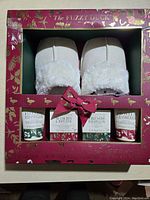 Front view of gift box showing white fuzzy slippers and four small jars/tubs labeled with scent names.