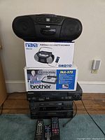 Overall stacked view of multiple vintage electronics including RCA boombox on top, Naxa boombox box, Brother fax machine box, Panasonic units below, and remote controls arranged in front.