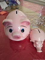 Front view of two pink ceramic piggy banks, large with detailed face and small with simple face, both on red surface.